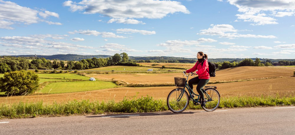 Kvinna cyklar på landsväg, blå himmel och sol i bakgrunden.